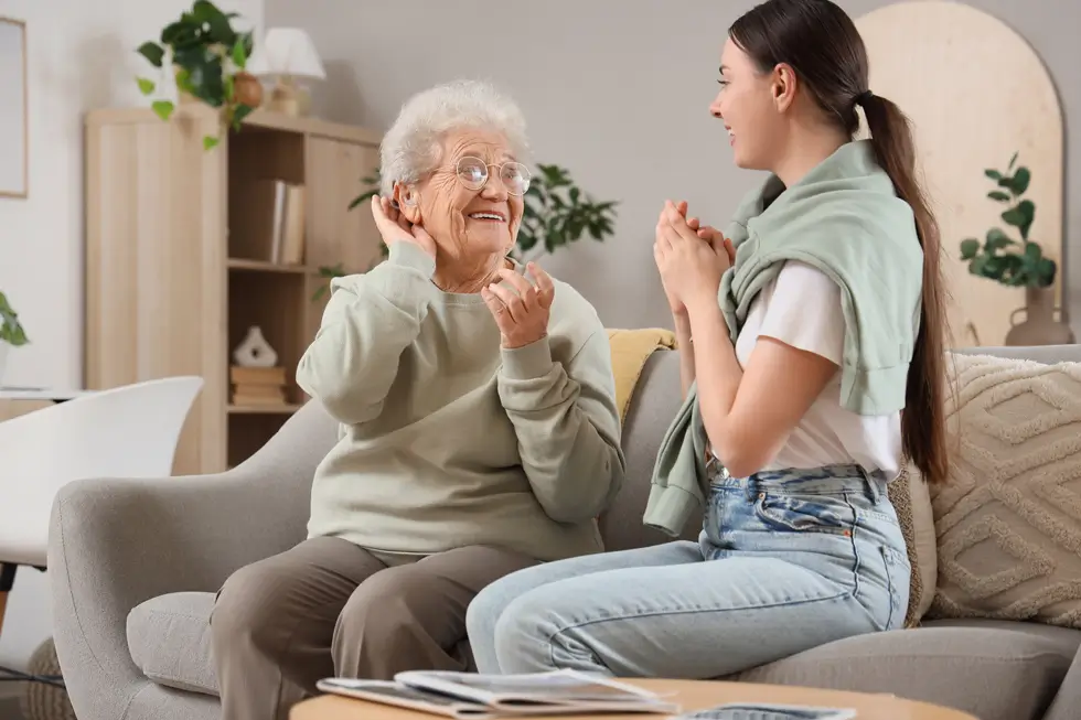 Zwei Frauen sitzen auf einem Sofa und reden, eine ältere mit grauen Haaren und Hörgerät und eine jüngere mit Pferdeschwanz
