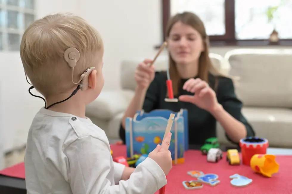Kleiner Junge mit Cochlea-Implantat am rechten Ohr hält Holzstab, sitzt an Tisch mit Spielzeug, Frau im Hintergrund mit Holzstab.