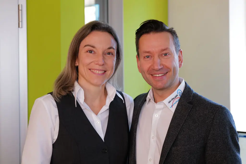 Zwei Personen stehen nebeneinander in einem Innenraum mit grünem Hintergrund, beide tragen Hemden, die linke Person zusätzlich eine schwarze Weste, die rechte einen dunklen Blazer